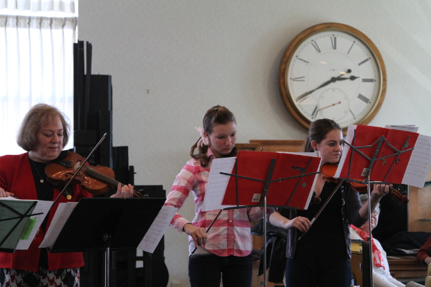 Drei Frauen auf der Bühne mit Violinen stehend und Musiknotenständer sowie Notenbücher vor sich; im Vordergrund sitzt ein Zuschauer; an der weißen Wand hängt eine Uhr, und im linken Eck ist ein weißer Vorhang zu sehen.