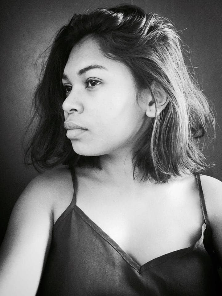 Eine selbstbewusste Frau in einem schwarzen Kleid posiert in einem Schwarz-Weiß-Foto, ihr glatter Bob und das dezente Make-up unterstreichen ihre selbstbewusste Haltung gegen eine Wand.