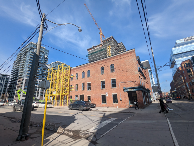 Eine belebte Stadtstraße mit fahrenden Autos, Fußgängern auf dem Gehweg, Strommasten mit Drähten, Straßenlaternen und Gebäuden, darunter ein teilweise errichtetes Hochhaus mit einem Kran vor dem bewölkten Himmel.