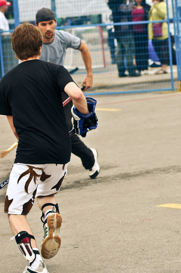 Zwei Menschen spielen Hockey auf einer Straße mit einem Zaun dahinter und einer Menge Menschen im Hintergrund.