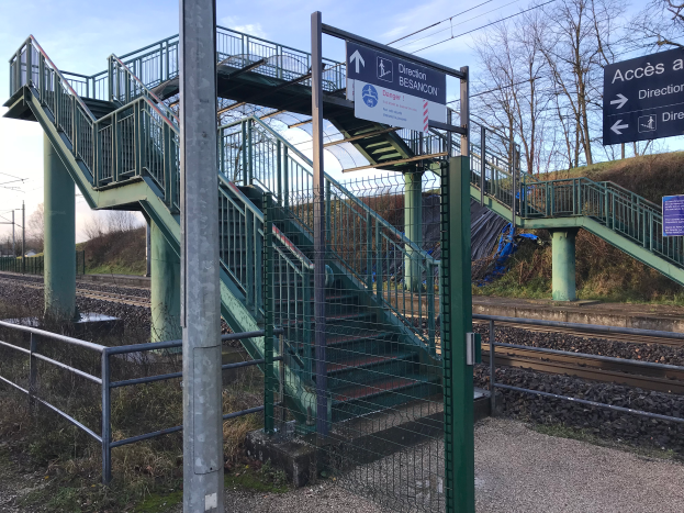 Ein Bahnhofsgebäude mit einer Treppe, die zu einem Bahnsteig führt, umgeben von Geländern und Pfählen, mit Texttafeln, Bäumen, Strommasten mit Drähten und einem bewölkten Himmel im Hintergrund.