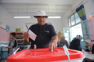 Ein Mann mit Hut stimmt bei einer Wahllokal ab, vor einer roten Wahlurne stehend, während andere sitzen und im Hintergrund schreiben, neben einem Fenster mit einem Vorhang.