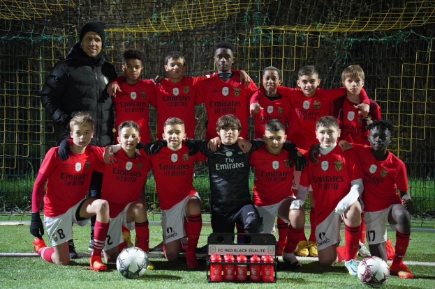 Eine Gruppe junger Jungs in roten T-Shirts und schwarzen Shorts posiert für ein Foto auf einem Fußballfeld, umgeben von Fußballbällen, einem Netz, einem Zaun und Bäumen im Hintergrund.