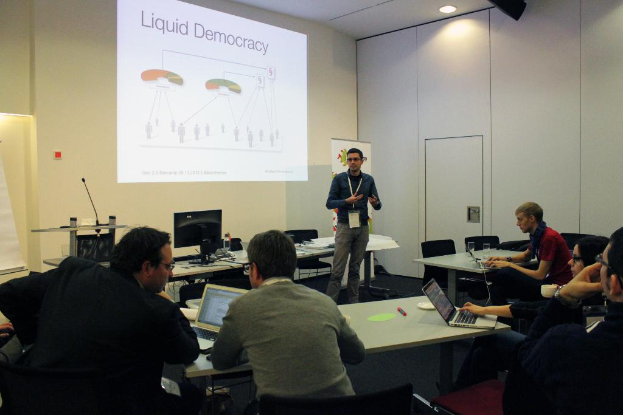 Fünf Personen in einem Konferenzsaal, die einem stehenden Mann gegenüber sitzen, mit Tischen, auf denen Laptops, Marker, Papiere, ein Monitor und ein Mikrofon sowie ein Projektionsschirm mit Text an der Wand zu sehen sind.