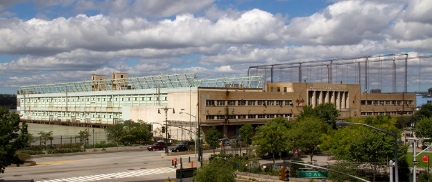 Eine StraГџe mit Fahrzeugen, BГ¤umen und StraГџenlaternen, die zu einem groГџen GebГ¤ude in der NГ¤he eines GewГ¤ssers fГјhrt.