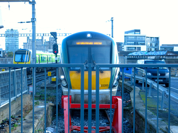 Zwei Züge auf parallelen Gleisen mit einem Zaun im Vordergrund, ein Auto auf der Straße rechts, eine Signalleuchte auf einem Pfahl, Gebäude im Hintergrund und ein Himmel über.