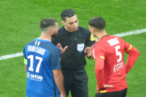 Ein Schiedsrichter in schwarzer Kleidung spricht mit zwei Männern auf einem Fußballfeld, einer in blauen und roten T-Shirts mit Aufschrift, beide tragen Brillen.