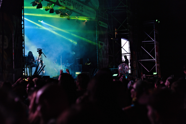 Menge bei einem Konzert mit einer Bühne, auf der Musiker spielen, die von Scheinwerfern beleuchtet wird, vor einem dunklen Hintergrund.
