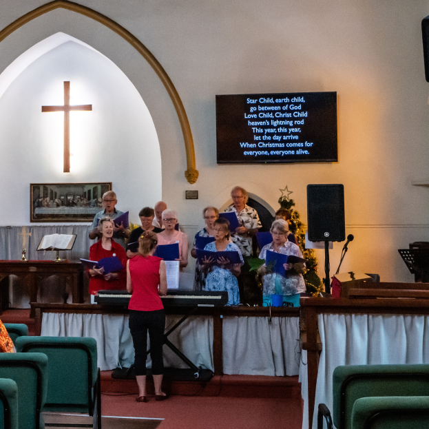 Eine Gruppe von Menschen vor einer Kirche, einige halten Bücher, Stühle links, Kreuzsymbol und Foto auf der Wand, ein Schild mit Text, ein Lautsprecher mit Ständer und ein Tisch mit einer Decke, scheinbar singend als Chor.