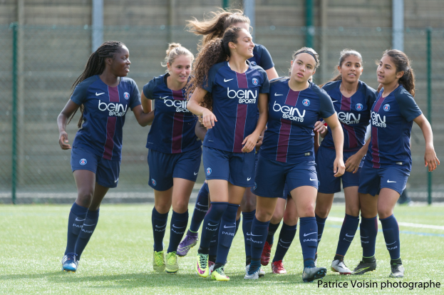 Gruppe junger Frauen beim Fußballspielen auf einem Rasenfeld mit Maschendrahtzaun und einer Wand im Hintergrund, mit dem Text "Paris Saint-Germain Women's Soccer" in der unteren rechten Ecke.