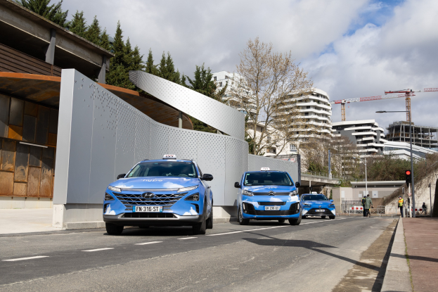 Zwei blaue autonome Autos fahren auf einer Straße neben einem Gebäude, mit Fußgängern auf dem rechten Gehweg und städtischen Elementen wie Bäumen, Gebäuden, Kränen und Verkehrszeichen unter einem klaren blauen Himmel.
