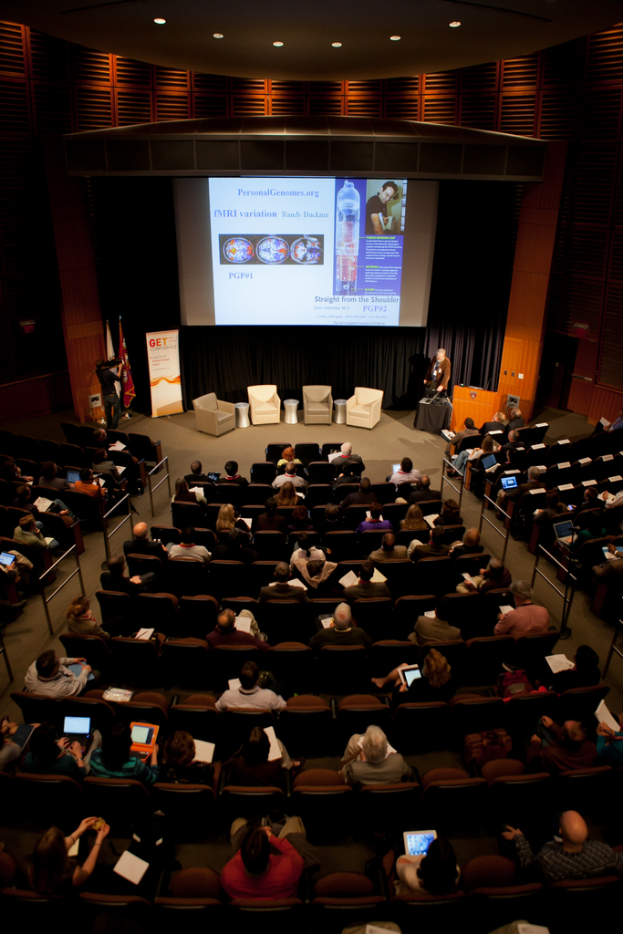 Ein mit sitzenden Menschen gefüllter Hörsaal, beleuchtet von zahlreichen Lichtern, mit Fahnen links, einem Podium rechts und einer Werbetafel links.