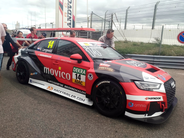 Audi RS3 DTM-Rennwagen auf einer Straße umgeben von Menschen, mit einem Zaun, Gras, einem Schild, einer Fahne, Pfosten und einem bewölkten Himmel im Hintergrund.