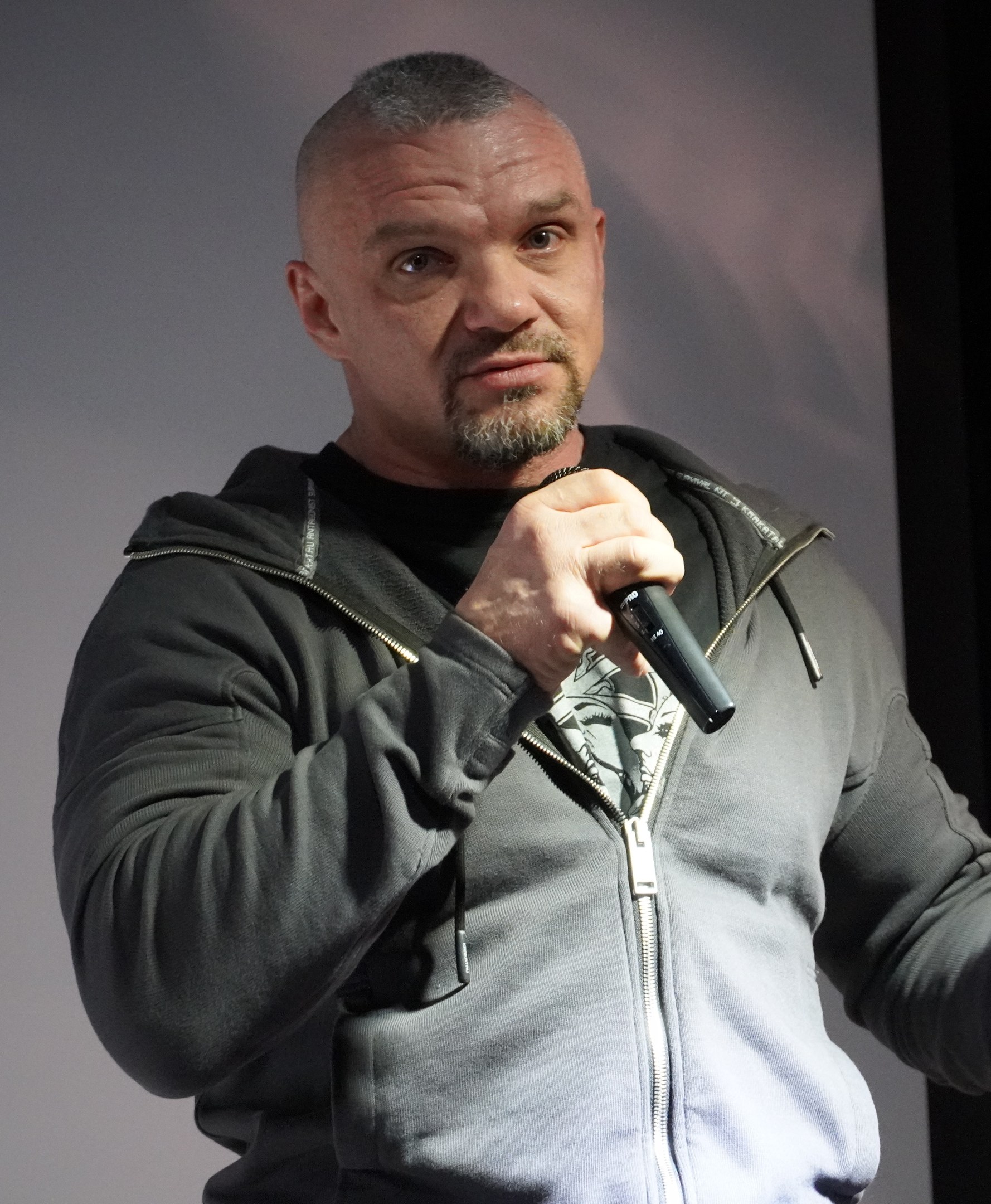 Ein Mann in einer schwarzen Kapuzenjacke und einem schwarzen Anzug mit einem weissen Hemd, der ein Mikrofon haltend vor einer Wand steht.