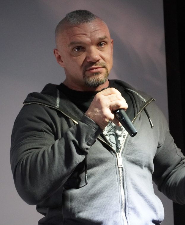 Ein Mann in einer schwarzen Kapuzenjacke und einem schwarzen Anzug mit einem weissen Hemd, der ein Mikrofon haltend vor einer Wand steht.