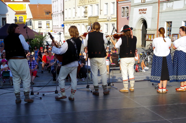 Vier Personen auf einer Bühne, die Geige spielen, mit einigen Zuschauern davor, Gebäuden im Hintergrund und zwei Frauen in der rechten Ecke.