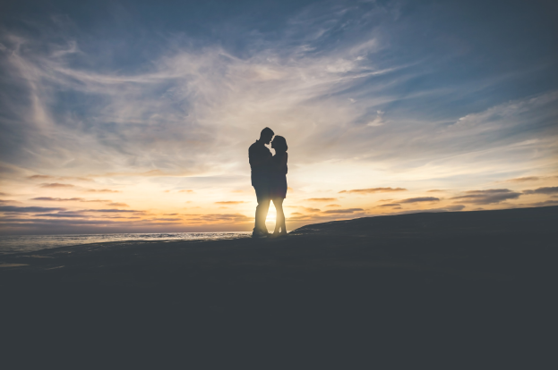 Silhouette eines Paares, das sich auf einem Strand bei Sonnenuntergang umarmt und küsst, mit leuchtenden orangefarbenen, pinken und violetten Himmel und tiefblauem Wasser im Hintergrund.