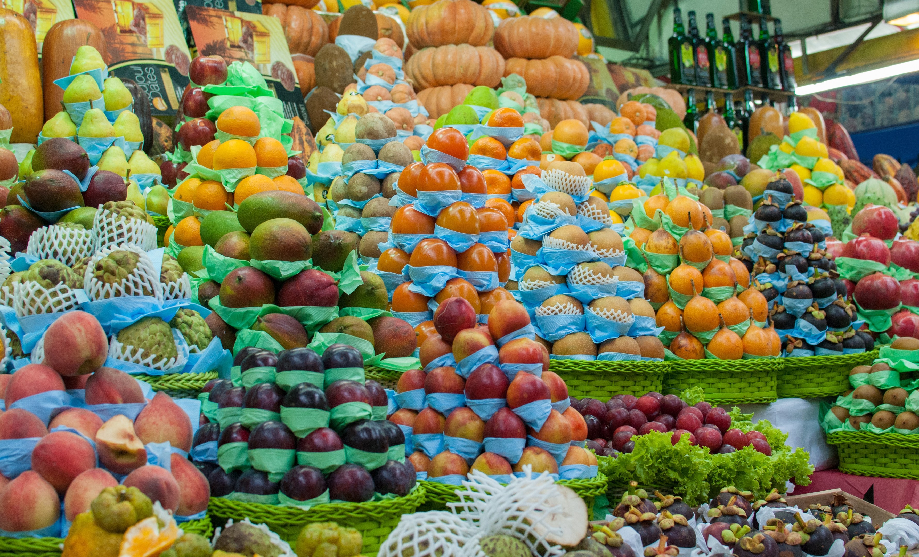 Ein Marktstand mit Äpfeln, Orangen, Birnen und anderen Früchten und Gemüsen sowie Flaschen auf Regalen und Deckenbeleuchtung.