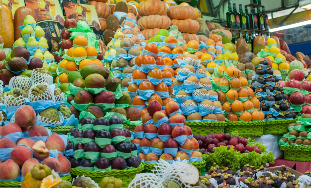 Ein Marktstand mit Äpfeln, Orangen, Birnen und anderen Früchten und Gemüsen sowie Flaschen auf Regalen und Deckenbeleuchtung.