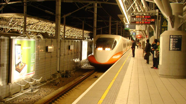 Eine Gruppe von Menschen steht auf einem Bahnsteig mit einem Zug auf den Schienen, einer Wand mit einer Tafel links und Pfählen mit Tafeln rechts.