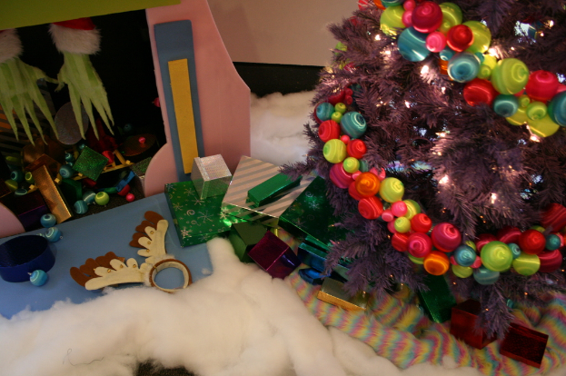 Drinnen steht rechts ein geschmückter Weihnachtsbaum mit Lichterkette und Schmuck, umgeben von Geschenkboxen und Dekorationsgegenständen, mit einer Hütte und Baumwolle oben links und Wolle unten.