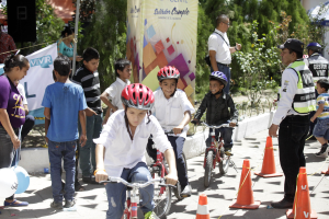 Kinder fahren Fahrräder auf einer Straße mit Verkehrskegeln, einige tragen Helme, andere stehen daneben, mit einem Banner, Bäumen und Gebäuden im Hintergrund.
