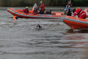 Eine Gruppe von Menschen in einem aufblasbaren Boot auf einem Fluss, mit zwei Personen im Wasser im Vordergrund und Pflanzen im Hintergrund, alle tragen Schwimmwesten und Helme, was auf einen Rettungseinsatz hinweist.