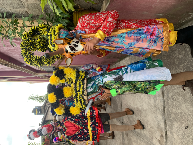 Eine Gruppe von Menschen in bunten Kostümen steht zusammen vor einem Gebäude, mit verschiedenen Gegenständen, Bäumen, Gebäuden und einem klaren blauen Himmel im Hintergrund während eines Karnevalsumzugs.