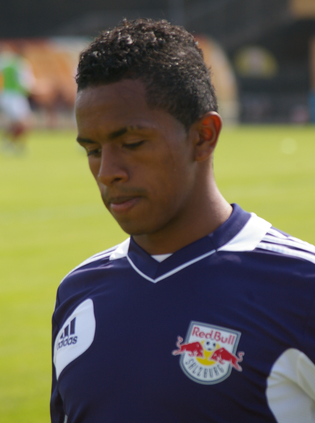 Ein junger Mann in einem blauen T-Shirt steht auf einem Fußballfeld, mit kurzen dunklen Haaren und einem entschlossenen Gesichtsausdruck, unscharfer Hintergrund.