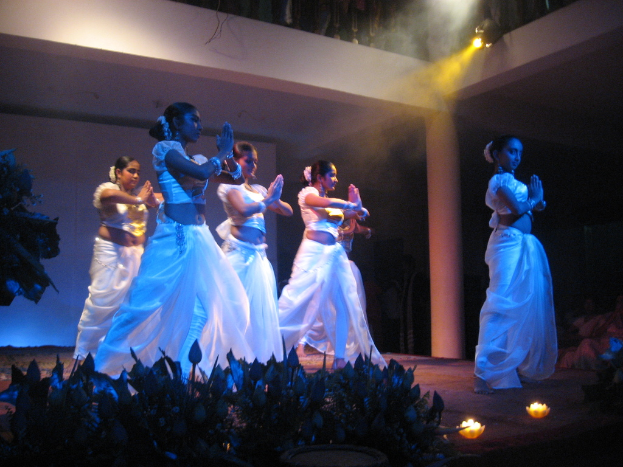 Eine Gruppe von Menschen tanzt auf einer Bühne mit Pflanzen davor und einem Publikum, das zusieht.