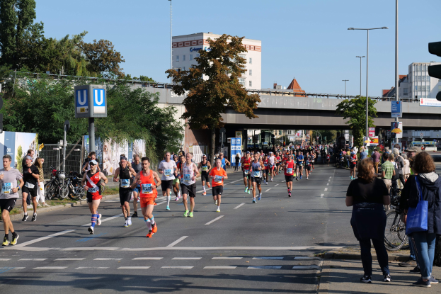 Gruppe von Menschen, die bei einem Marathon auf einer Straße mit Bäumen, Laternen, Schildern und einem Zaun laufen, mit Fahrrädern in der Nähe und einer Brücke im Hintergrund unter einem klaren blauen Himmel.