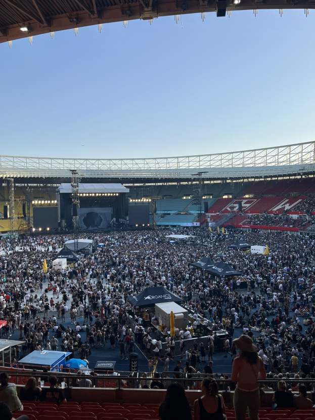 Große Menge sitzend und stehend in einer Halle mit Geländern, Zelten und Deckenbeleuchtung, vor einem sichtbaren Himmel als Hintergrund, was auf ein Konzert in der O2 Arena in London hindeutet.