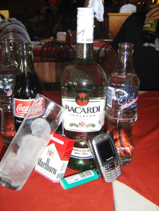 Eine Sammlung von Flaschen, darunter Alkohol, Wasser und eine Coca-Cola, ein Glas mit Eiswürfeln, ein Feuerzeug und ein Mobiltelefon auf einem Tisch mit Stühlen und Menschen, die Wein im Hintergrund trinken.