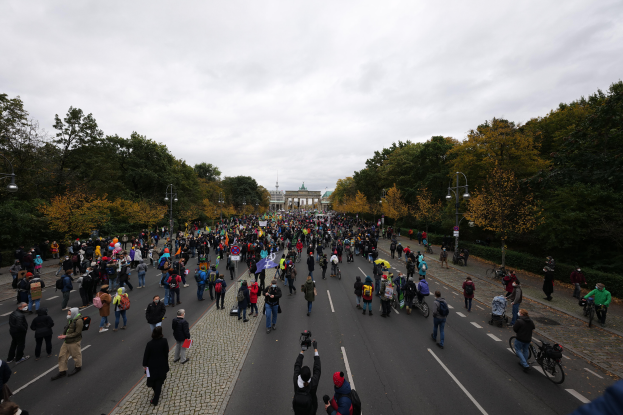 Eine große Gruppe von Menschen, die eine von Bäumen gesäumte Straße in Berlin entlanggeht, einige halten Kameras, mit einem Gebäude im Hintergrund und einem klaren Himmel darüber.