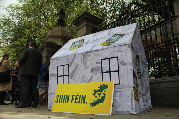 Ein Zelt mit einem Werbeplakat davor, umgeben von einer Menge, mit einem Eisenzaun, Säulen, Straßenlaternen, Bäumen und Himmel im Hintergrund.