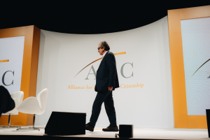 Ein Mann im Anzug und Krawatte geht auf einer Bühne vorbei an Lautsprechern, Stühlen und einem Banner mit Text.