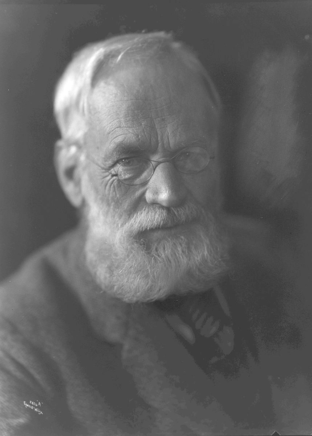 Ein ernster aussehender älterer Mann mit Vollbart und Brille, der konzentriert in die Ferne schaut, vor dunklem Hintergrund.