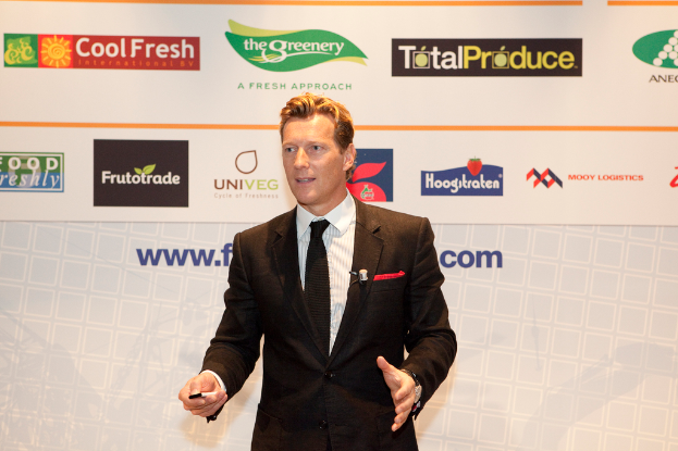 Ein Mann in einer schwarzen Blazer steht und hält ein Objekt, spricht, mit einem weißen Banner und einer weißen Tafel mit organisationalen Logos im Hintergrund.