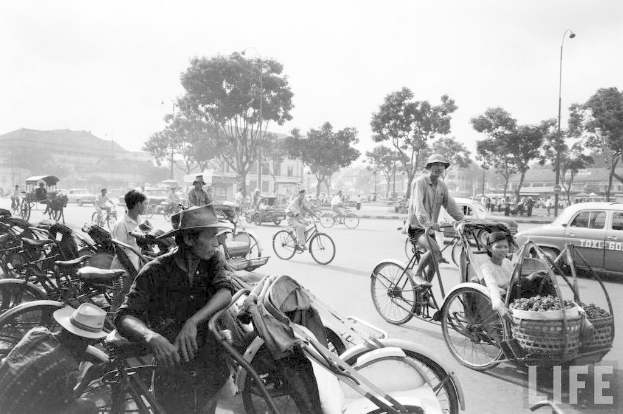 Eine belebte Straße mit Fahrrädern, Autos und anderen Fahrzeugen, die vorbeifahren, mit Bäumen und Gebäuden im Hintergrund.
