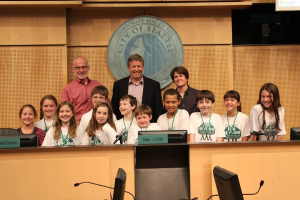 Gruppe von Kindern und Erwachsenen, die vor einem Rednerpult auf einer Stadtverordnetenversammlung lächeln, mit Monitoren und Mikrofonen auf einem Tisch im Vordergrund, einem Logo an der Wand und einem Bildschirm auf der rechten Seite.