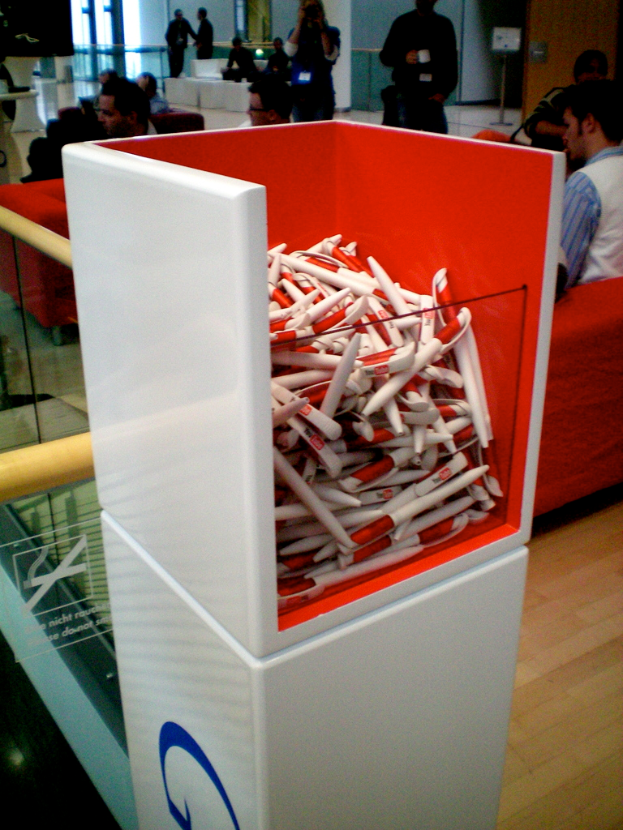 Ein Kasten mit Stiften im Vordergrund, mit Gruppen von Menschen im Hintergrund, die sitzen und stehen, ein Schild an einem Pfahl, eine Wand, ein Holzboden und Text auf einem Spiegel.