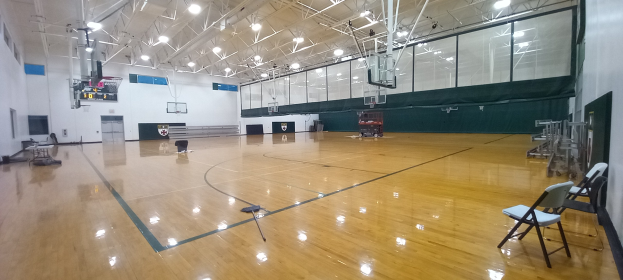 Großer Turnsaal mit Holzboden, Stühle an den Wänden, ein Basketballfeld und Deckenbeleuchtung.