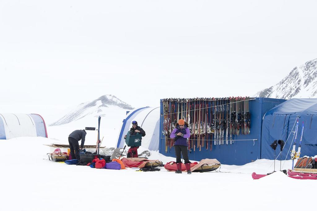 Drei Personen stehen auf einer schneebedeckten Landschaft mit verstreuten Taschen, Zelt mit Skiern dahinter und schneebedeckten Hügeln im Hintergrund unter einem klaren Himmel.