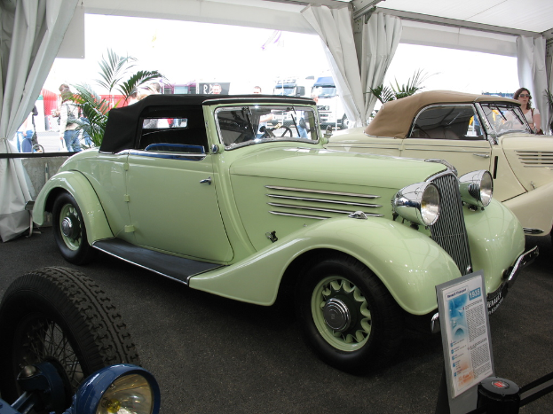 Mehrere Oldtimer stehen auf einem Boden mit Vorhängen und Pflanzen im Hintergrund, während weiter hinten auf der Straße einige Menschen und Fahrzeuge zu sehen sind.