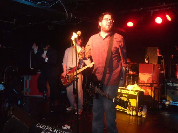 Drei Personen auf einer Bühne mit Mikrofonen und Scheinwerfern, jede hält eine Gitarre.