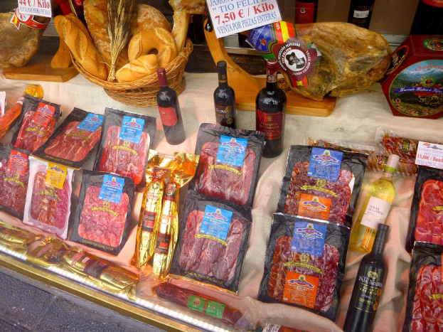 Eine Theke eines Metzgerladens mit verschiedenen Fleischsorten, Flaschen, einem Korb und anderen Lebensmitteln sowie daneben Texttafeln.