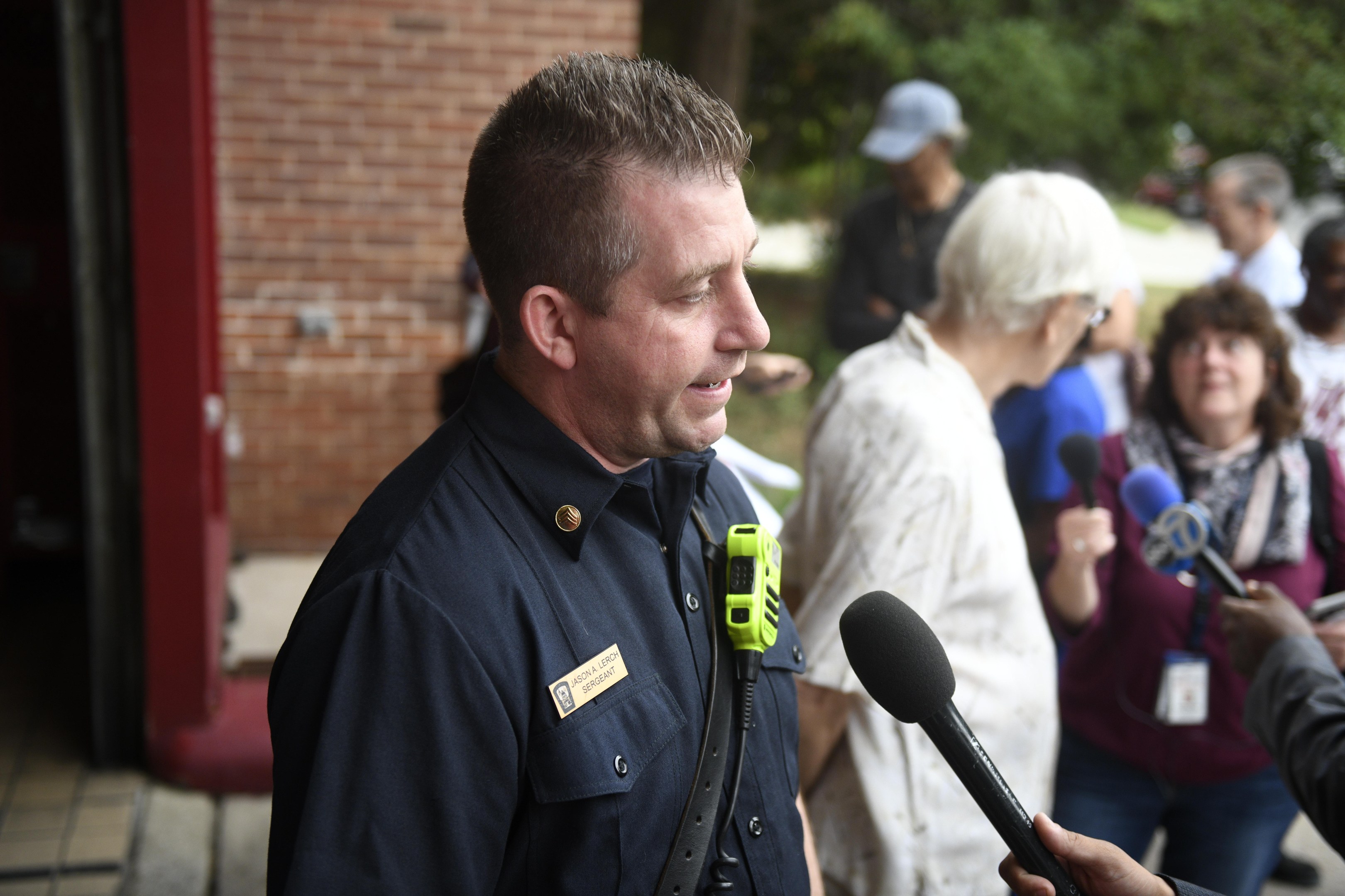 Ein Polizeibeamter, der zu einer Gruppe von Menschen spricht, mit Mikrofonen, Bäumen und einem Gebäude im Hintergrund.
