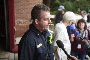 Ein Polizeibeamter, der zu einer Gruppe von Menschen spricht, mit Mikrofonen, Bäumen und einem Gebäude im Hintergrund.